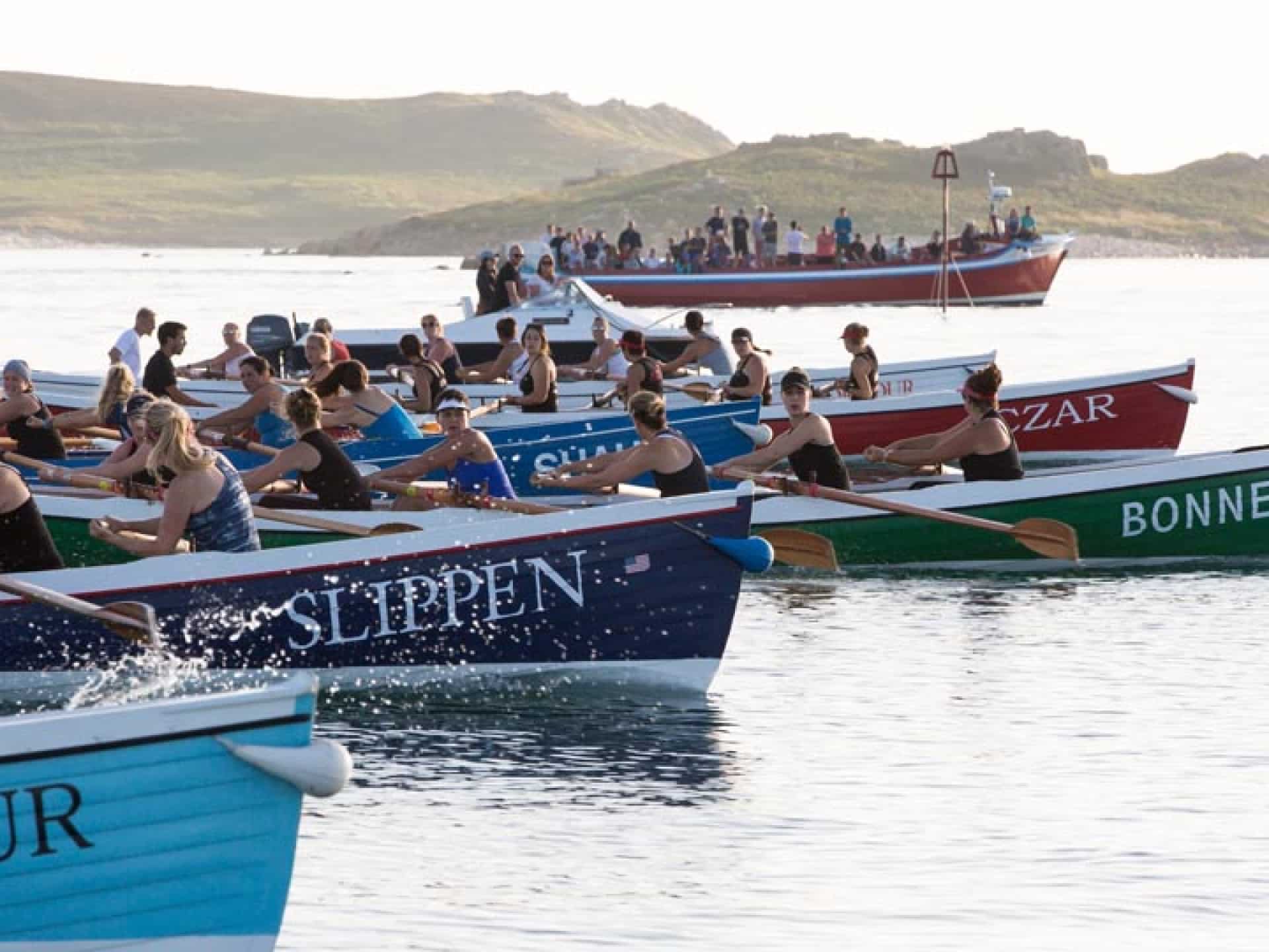 Rowers compete in several colorful boats on calm water, with spectators watching from a larger boat in the background and a hilly shoreline visible under bright daylight.