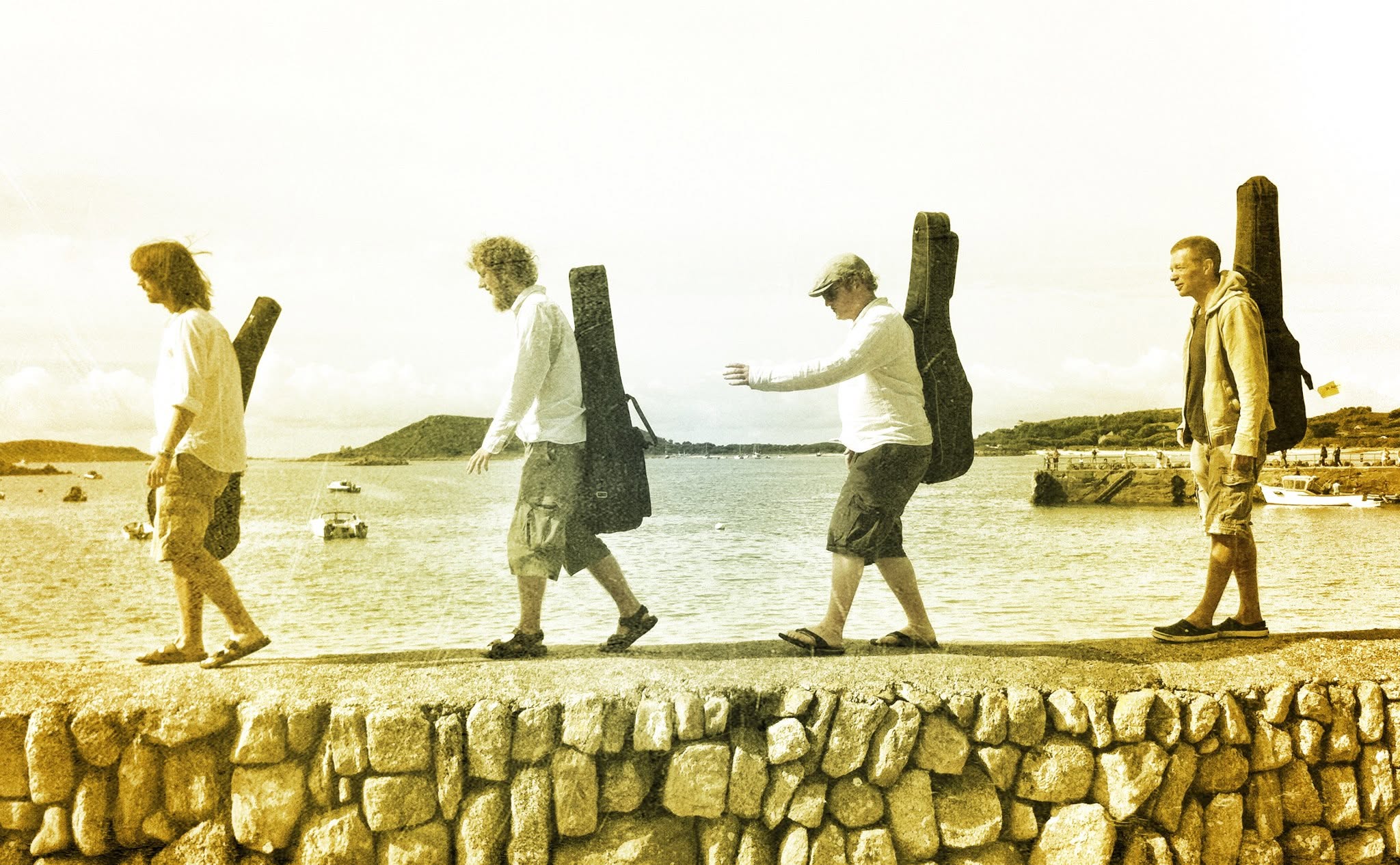 Four people with guitar cases walk in a line along a stone wall by the sea, mimicking the iconic Abbey Road pose. The photo has a warm, vintage filter.
